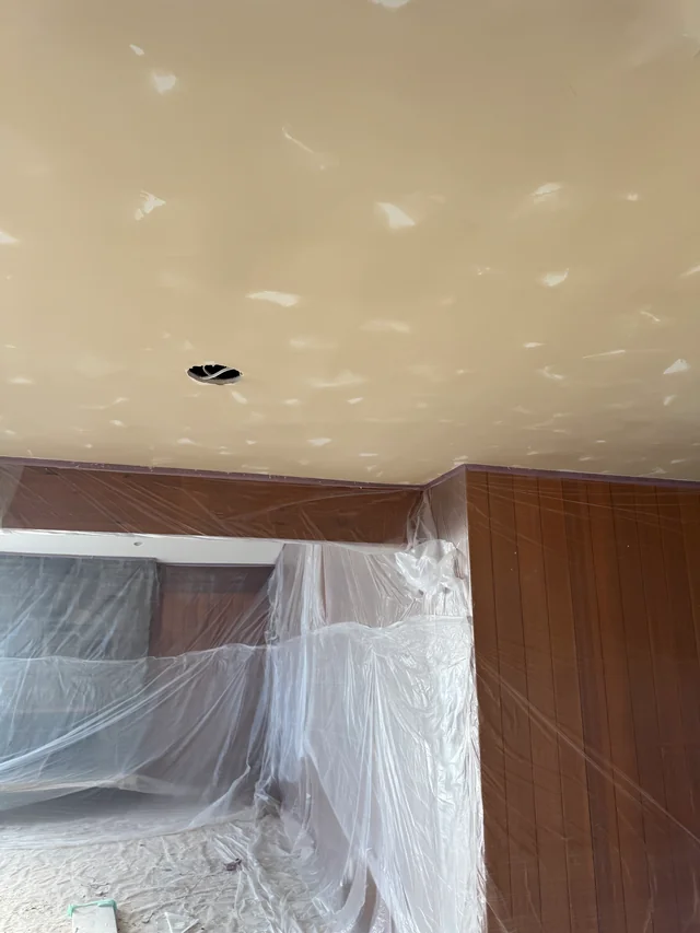 Joint compound across a ceiling beside wood paneling and plastic sheeting protecting the adjoining area.