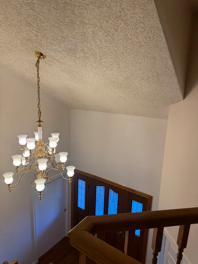 Finished textured ceiling and smooth walls above a stairwell with an uncovered chandelier and wood railing.