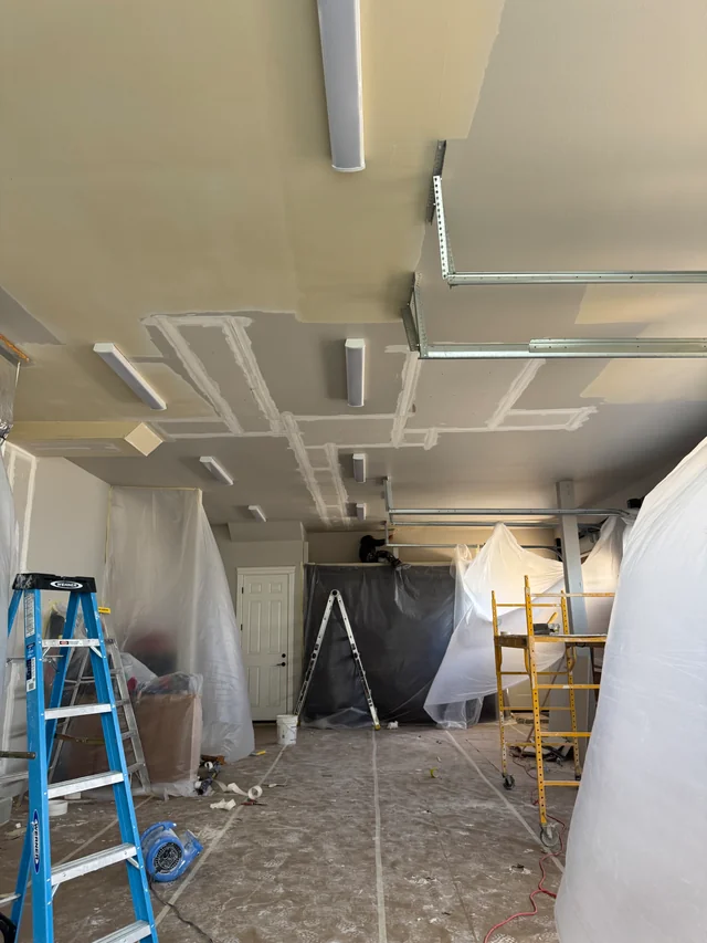Drywall ceiling patches and taped joints in a garage, with ladders and plastic protecting the surrounding work area.