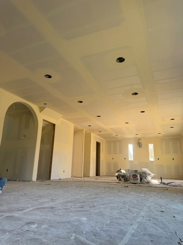 Taped drywall joints across an open room with ceiling light openings, arched doorways and covered floors.