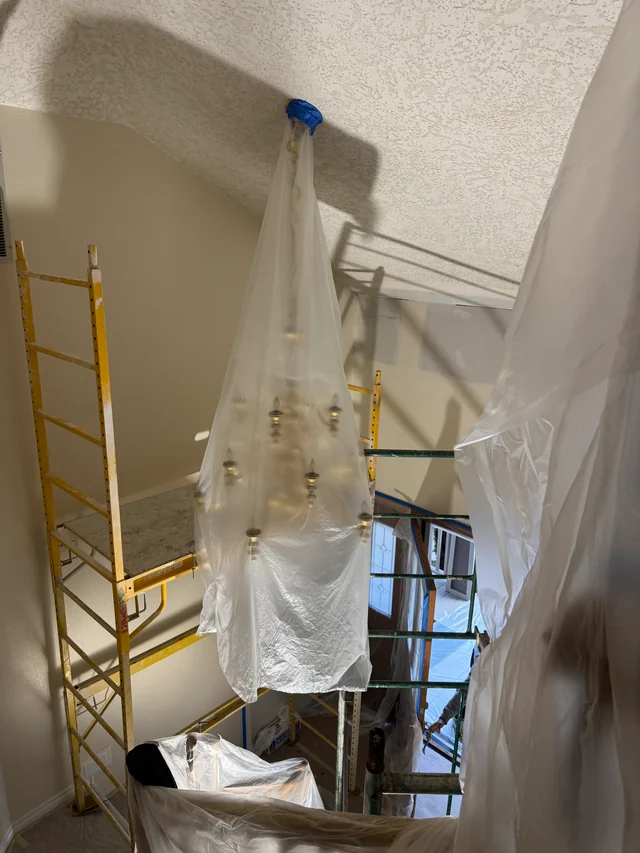 A chandelier wrapped in clear plastic beside scaffolding during drywall work in a tall stairwell.