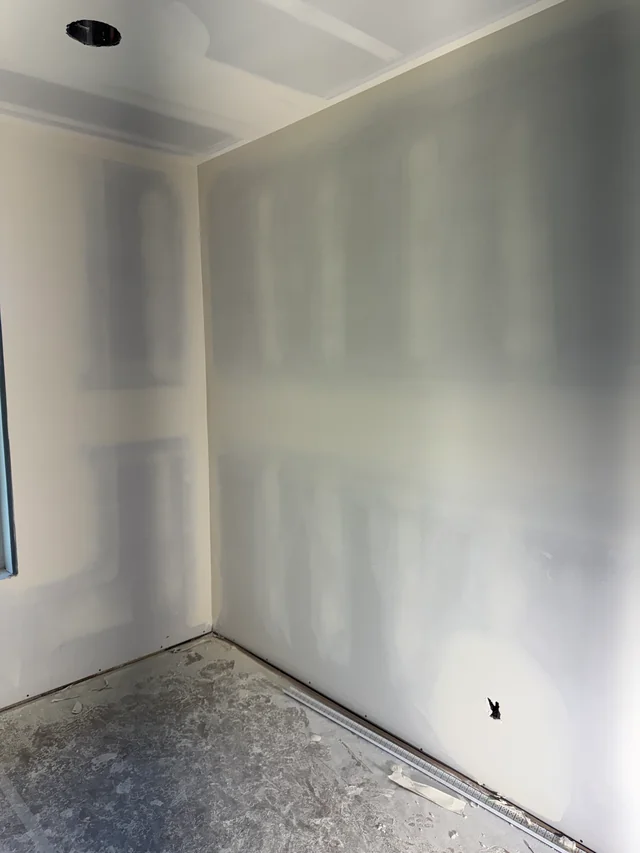 Taped drywall seams across a room corner and ceiling beside a window, with protective covering on the floor.