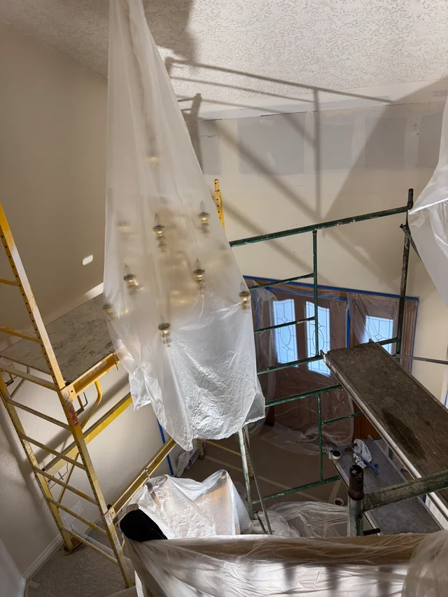 Scaffolding spans a stairwell beneath a textured ceiling while the chandelier is protected with clear plastic.