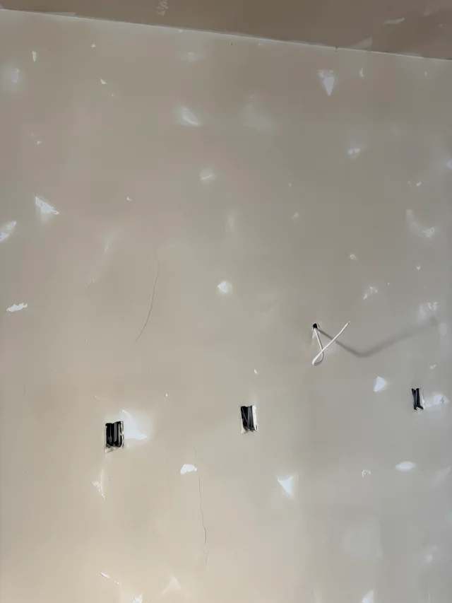 Close view of a drywall wall with joint compound over fasteners around electrical openings and loose wiring.