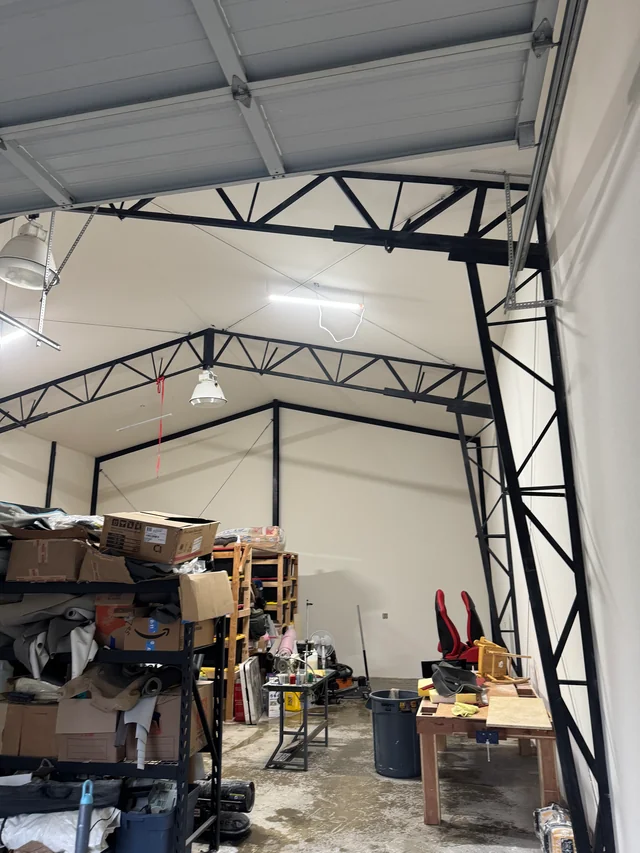 Drywall ceiling joints in a workshop with exposed black steel trusses, garage door tracks and equipment below.