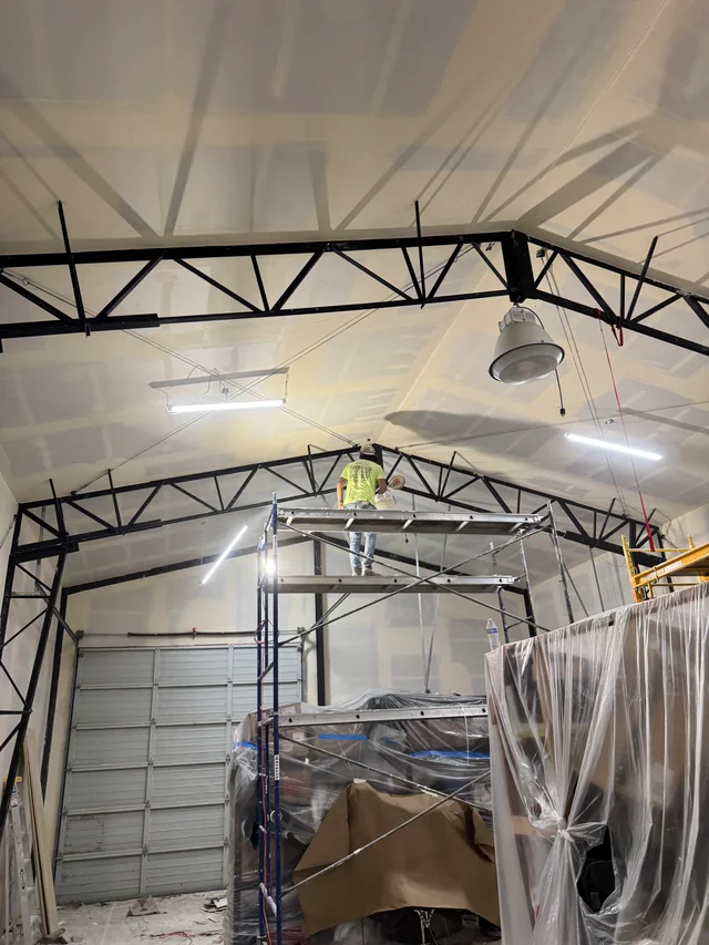 Taped drywall ceiling around black steel trusses above scaffolding and plastic-covered workshop equipment.
