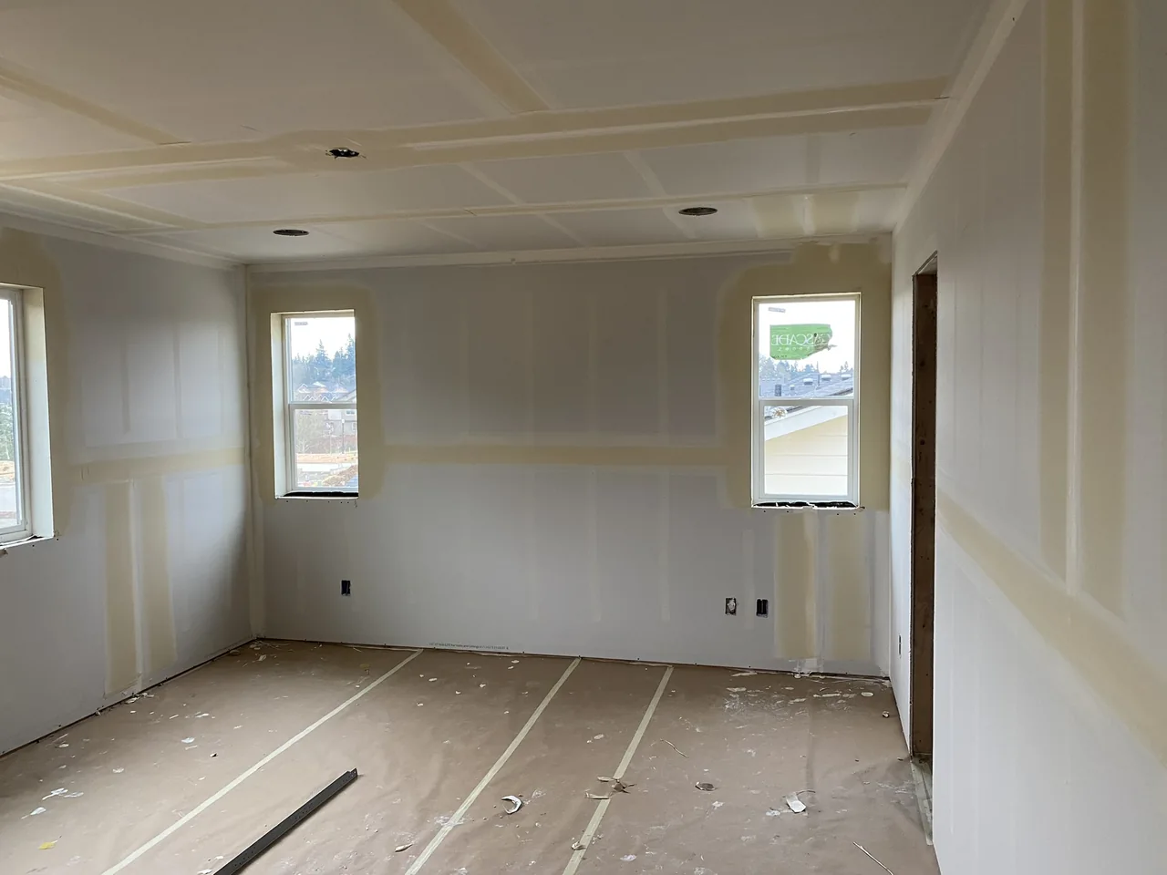 Drywall finishing with taped joints across the walls and ceiling of a bright room.